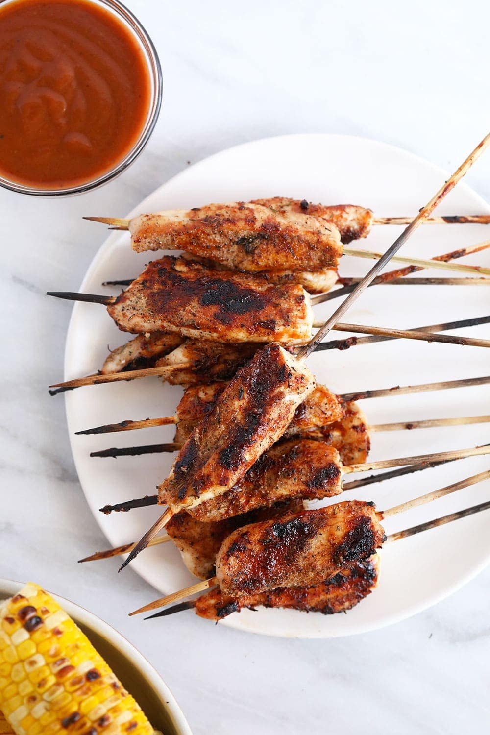 Grilled skewers on plate next to bbq sauce and grilled corn. 