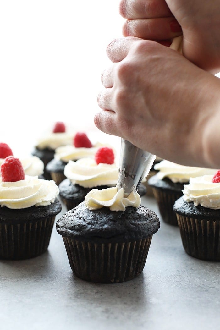 healthier chocolate cupcakes being piped with vanilla frosting and topped with a fresh raspberry