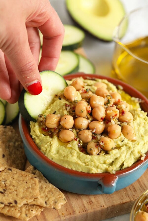 a person dipping a cucumber into a bowl of avocado hummus.