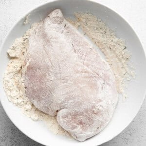 A raw chicken breast covered in flour on a white plate on a white surface.