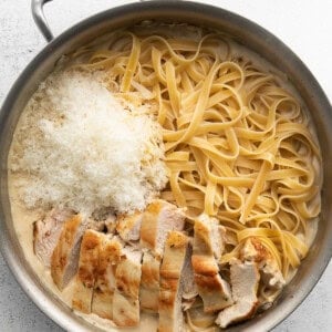 A stainless steel pan contains cooked fettuccine pasta, sliced grilled chicken breast, and grated Parmesan cheese, with a creamy sauce.