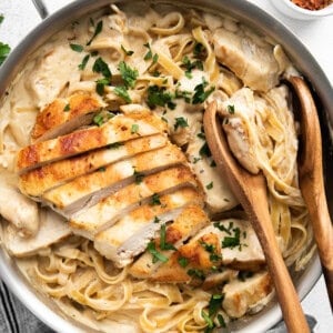 A skillet filled with creamy fettuccine Alfredo topped with sliced grilled chicken and garnished with chopped parsley. Wooden utensils are placed on the side.