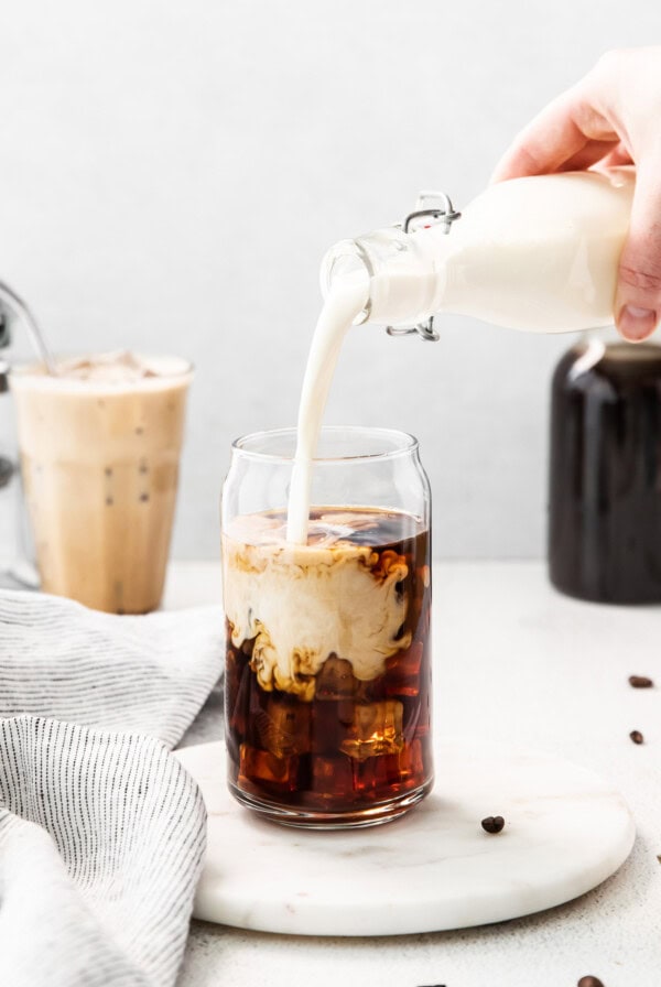 a person pouring milk into a glass of iced coffee.