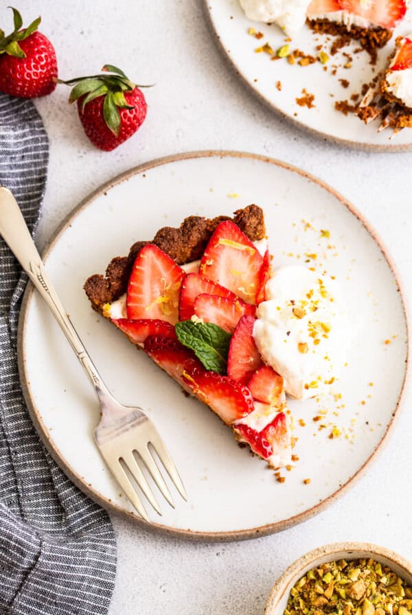 A slice of strawberry tart with whipped cream and pistachios on a plate.