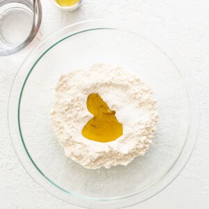 A bowl of flour, eggs and oil on a white surface.