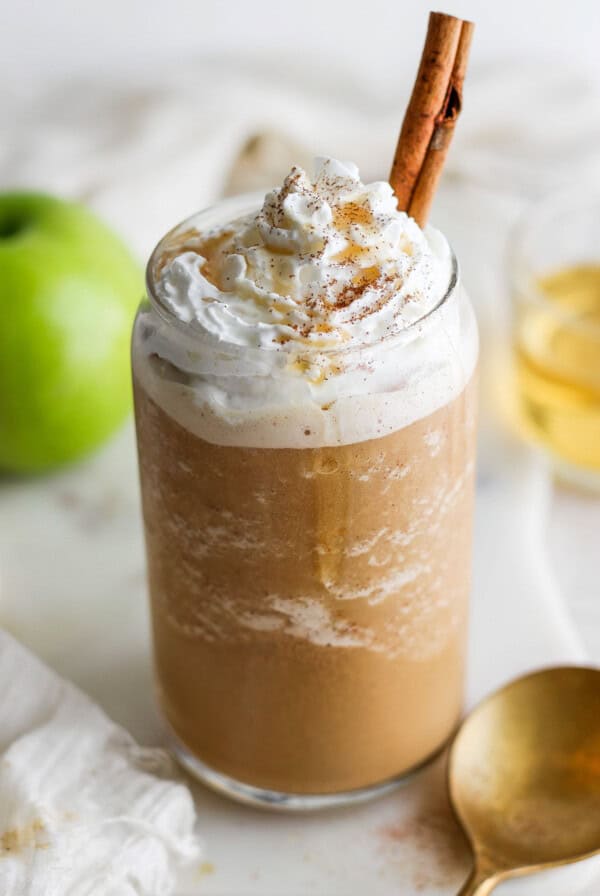 A mug with whipped cream, cinnamon sticks and apples.