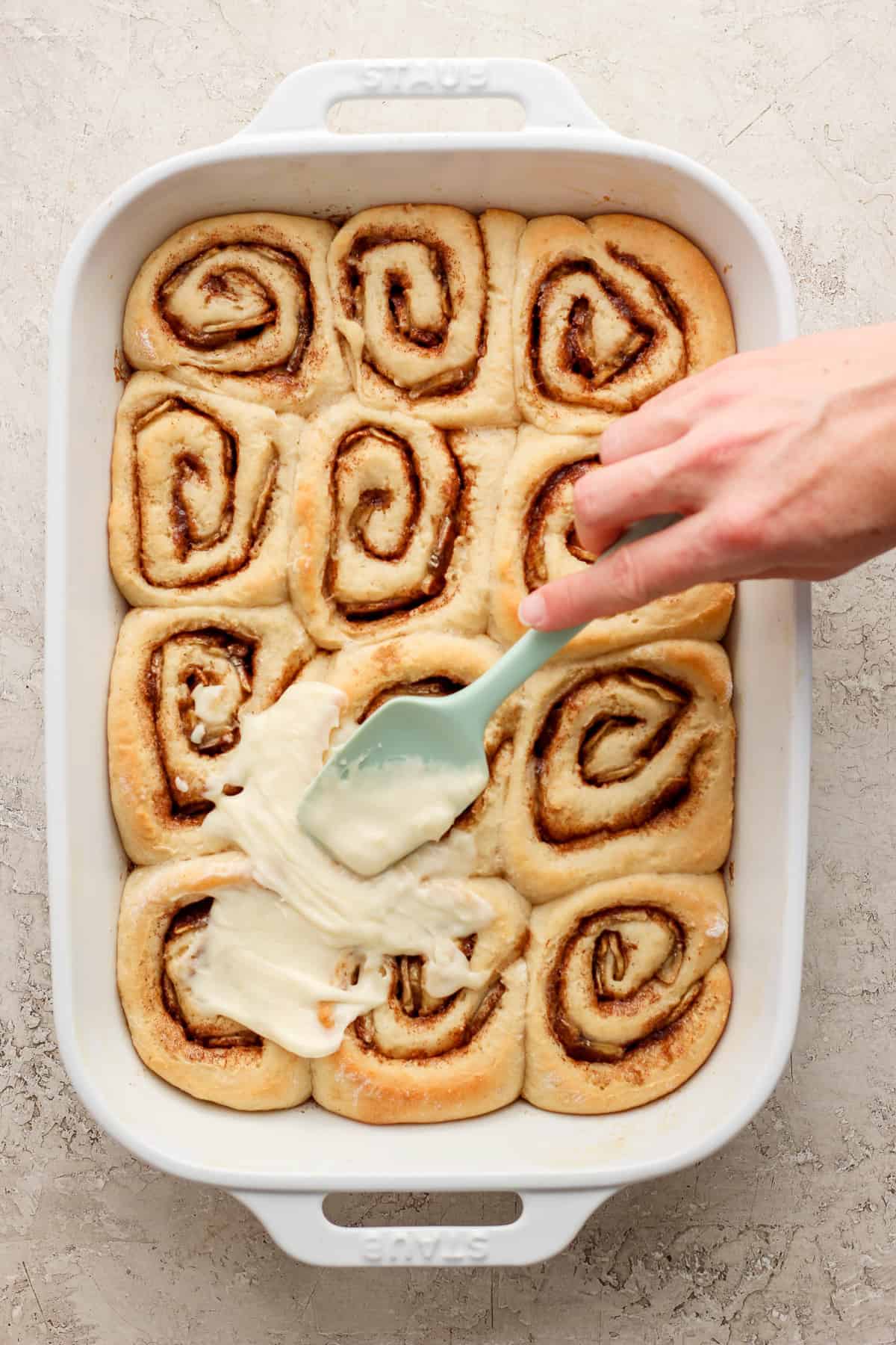 A person is dipping a spatula into a dish of cinnamon rolls.