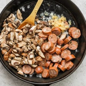 Sausages and mushrooms in a skillet with a wooden spoon.