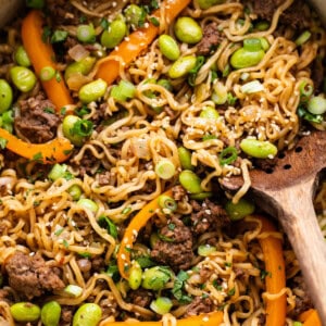 A pan full of noodles and vegetables with a wooden spoon.