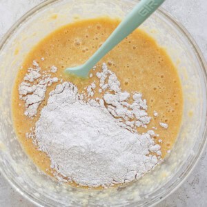 A glass bowl containing a mixture of flour and a liquid batter with a green silicone spatula.