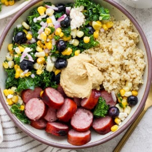 A bowl of quinoa, sliced sausage, and mixed vegetables, topped with a dollop of hummus.