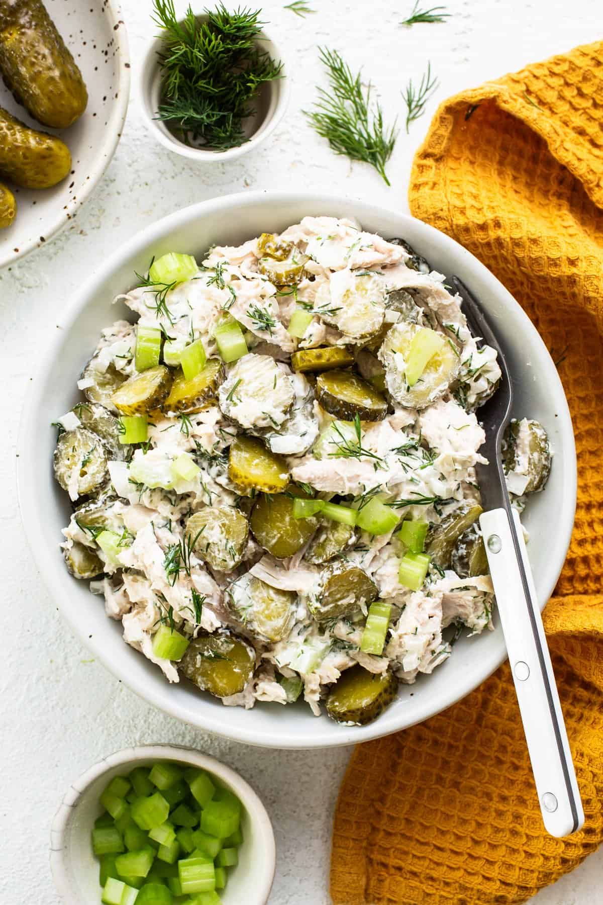 A bowl of chicken and pickle salad garnished with dill, accompanied by a serving fork, on a white surface with pickles and chopped celery nearby.