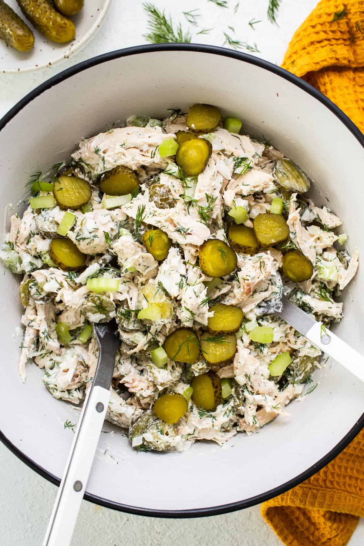 A bowl of dill pickle chicken salad with sliced pickles and fresh dill, accompanied by serving utensils.