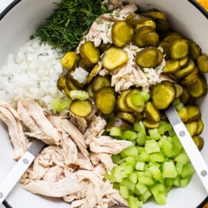 Ingredients for a recipe including shredded chicken, chopped onions, sliced pickles, and diced celery in a bowl with tongs.