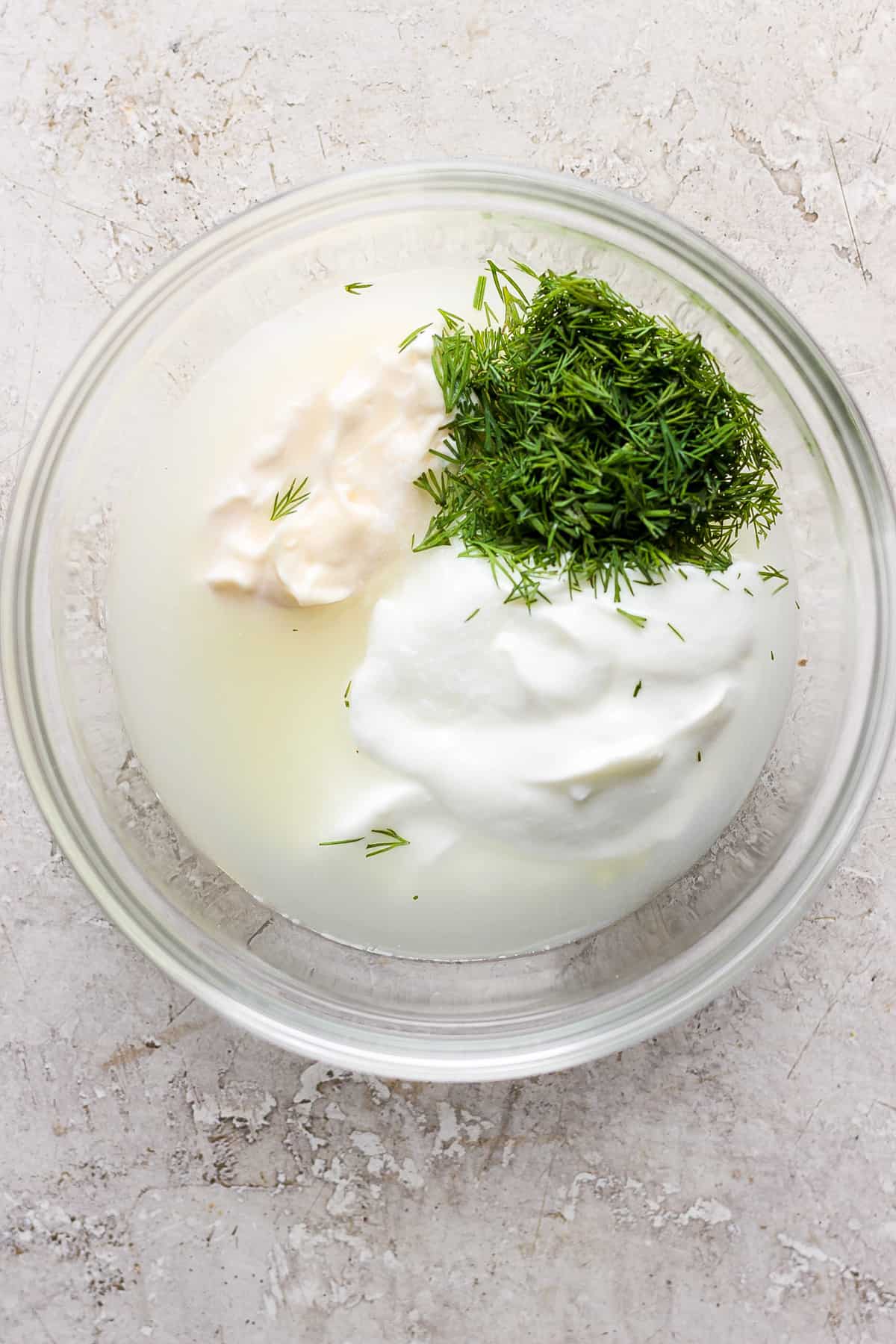 A glass bowl containing sour cream, dill, and a liquid, possibly part of a recipe preparation.