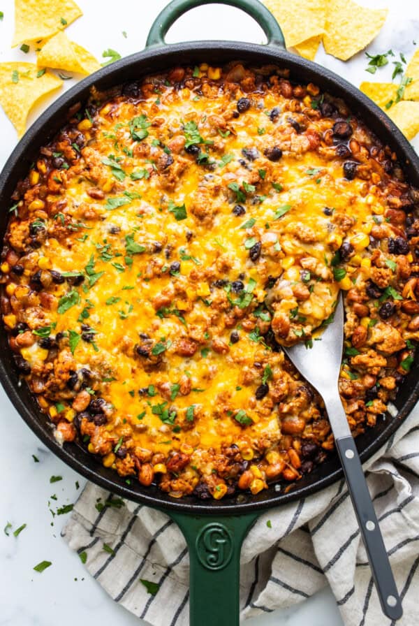 A skillet with a baked cheesy bean and ground meat casserole garnished with herbs, accompanied by tortilla chips.