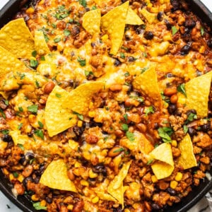 A skillet of baked nachos with melted cheese, ground meat, beans, and corn, garnished with crispy tortilla chips.