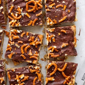 Chocolate-covered toffee bars topped with pretzel pieces on parchment paper, cut into rectangles, with a knife resting to the side.