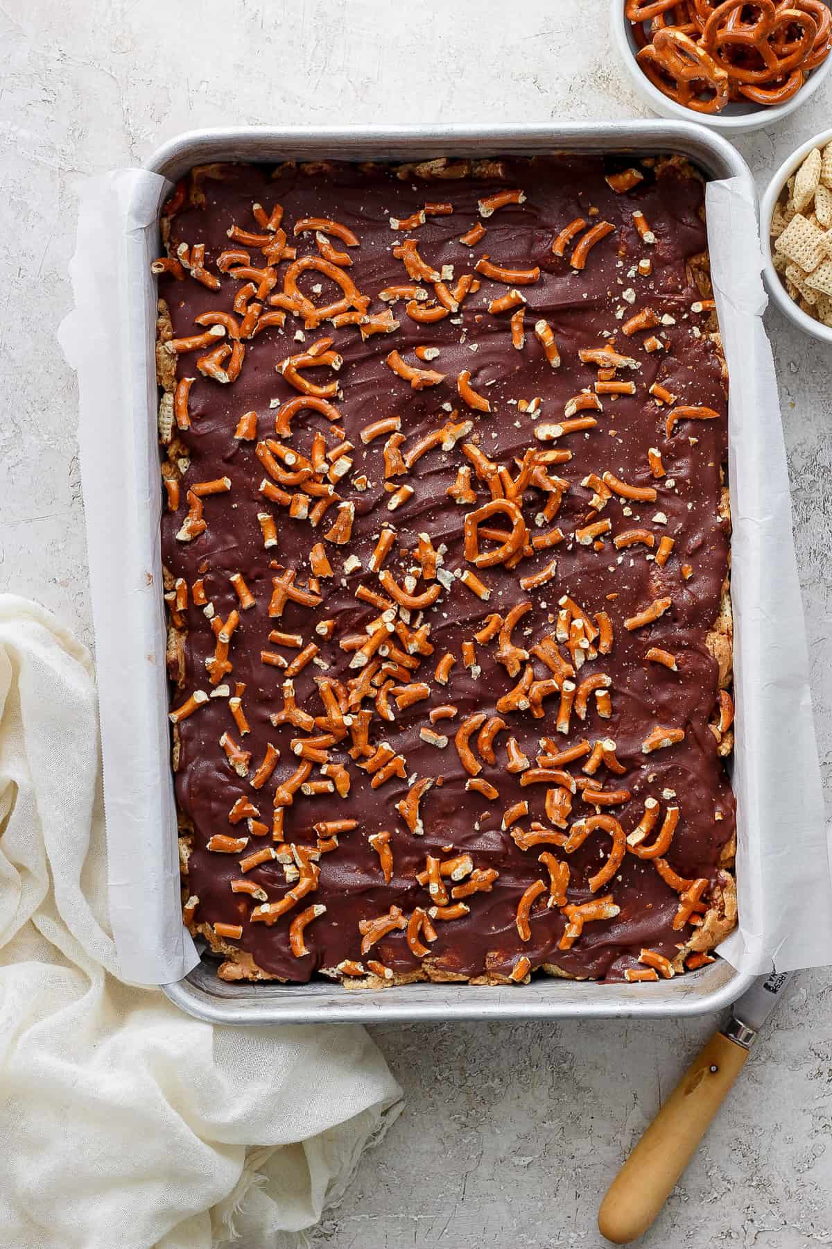 A tray of chocolate-covered toffee sprinkled with pretzel pieces on a kitchen counter, accompanied by a cloth and a spatula.