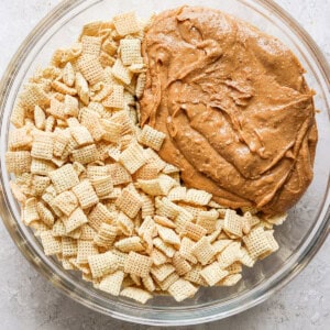 A glass bowl containing a mixture of cereal pieces and a large dollop of peanut butter on a light surface.