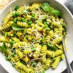 A bowl of pesto pasta garnished with peas, grated cheese, and basil leaves.