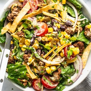 A vibrant taco salad with ground meat, mixed greens, tomatoes, cheese, corn, black beans, and crispy tortilla strips, served with a creamy dressing on the side.