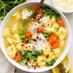 A bowl of chicken noodle soup garnished with parsley, shredded cheese, and a sprinkle of red pepper flakes, served with lemon wedges on the side.