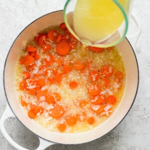 Pouring chicken broth into a pot with cooking onions and carrots.