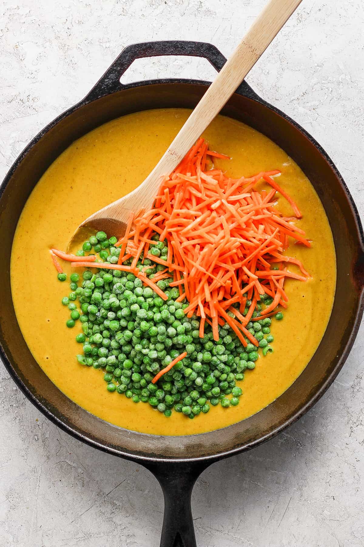 A cast iron skillet containing creamy soup garnished with a spoonful of green peas and a scattering of grated carrots on a light-colored surface.