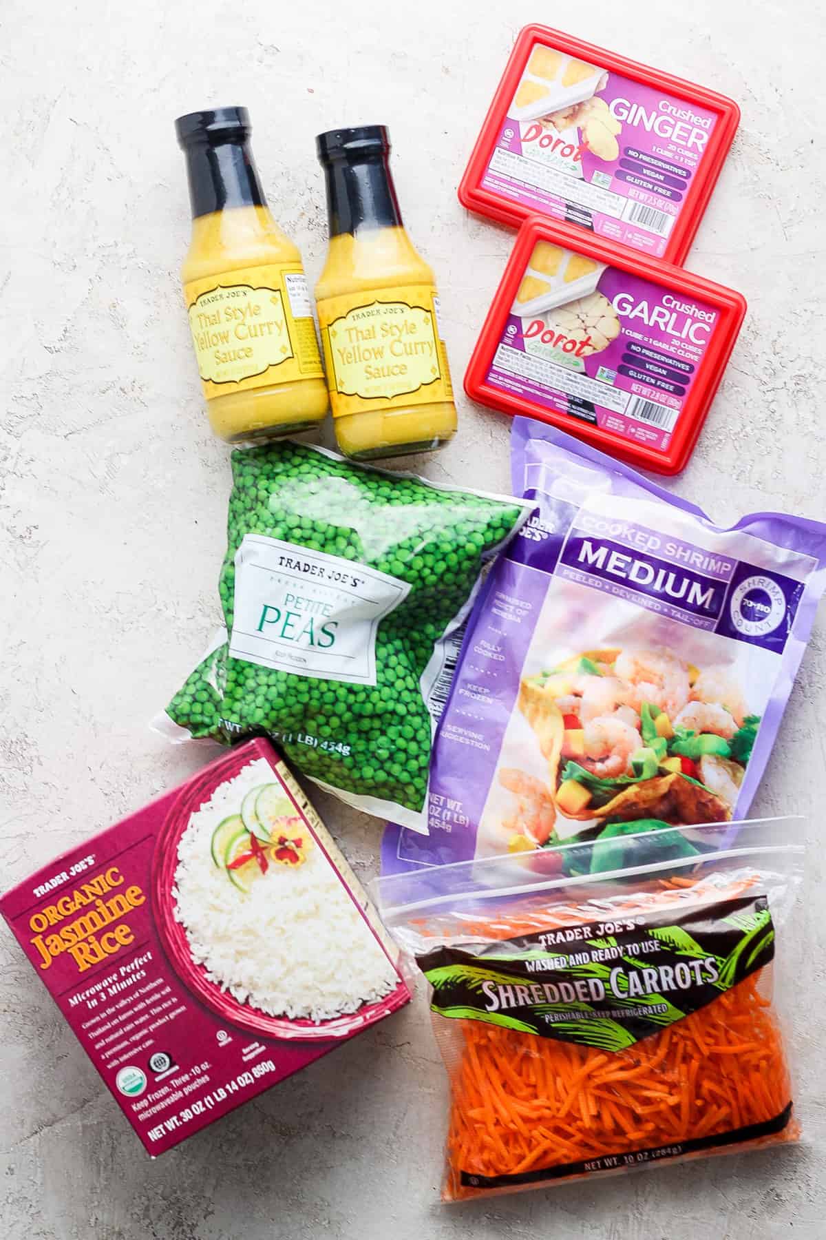 A collection of various food items, including bottles of thai yellow curry sauce, a box of ginger, packaged garlic, organic jasmine rice, frozen peas, and shredded carrots, arranged on a light textured surface.