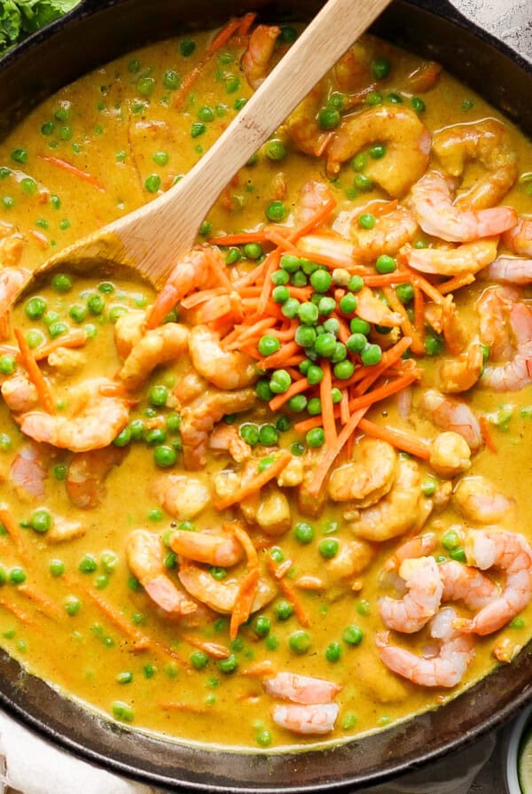 A wooden spoon is stirred into a skillet containing a shrimp curry with green peas and shredded carrots, accompanied by a side of white rice and lime wedges.