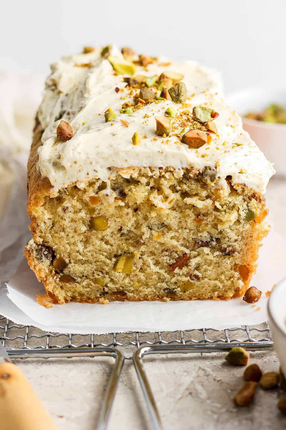 A loaf of frosted cake with nuts and chopped pistachios on top, partially sliced, displayed on a piece of parchment paper.