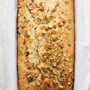 A freshly-baked loaf of bread with pistachios sprinkled on top, sitting in a metal baking pan lined with parchment paper.