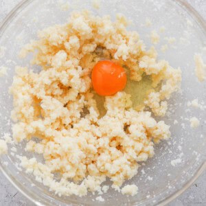 A glass bowl containing crumbled dough and a whole raw egg on a textured surface.