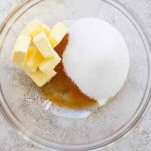 Ingredients for baking, including butter, white sugar, and brown sugar, are placed in a glass mixing bowl on a textured surface.