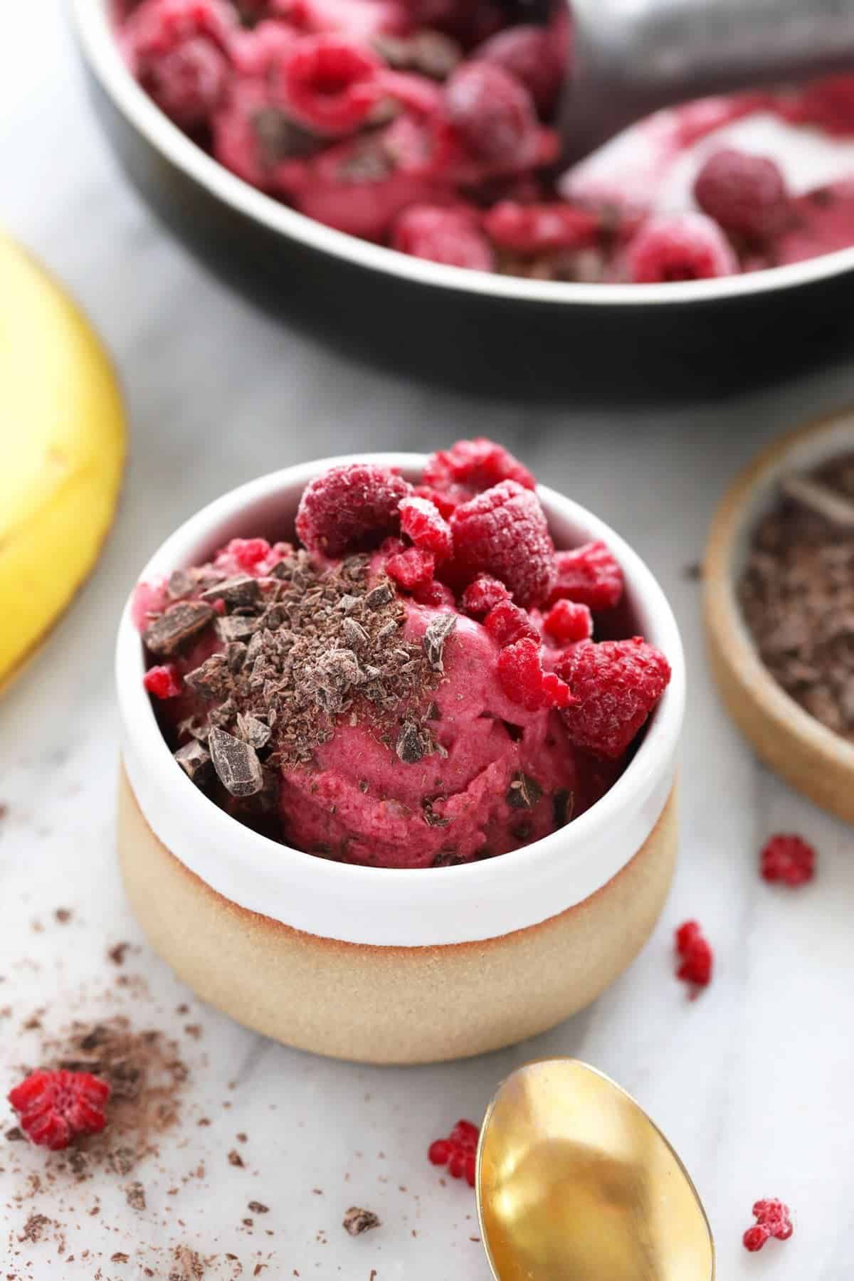 A bowl of raspberry nice cream topped with chocolate shavings and whole raspberries, with a banana and spoon nearby on a marble surface.