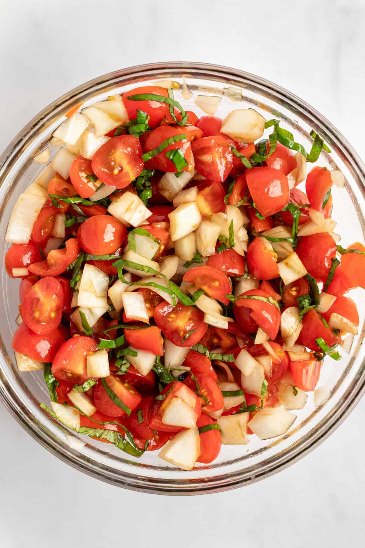 A glass bowl filled with a fresh salad of chopped tomatoes, cucumbers, and herbs on a white surface.