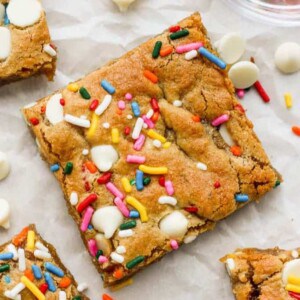 A close-up of a square Funfetti blondie topped with colorful sprinkles and white chocolate chips, with more scattered around it.