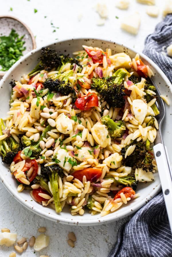 A bowl of orzo salad with roasted broccoli, tomatoes, and pine nuts, sprinkled with cheese and herbs, on a textured white surface.