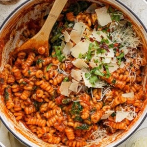 A pot of tomato-based pasta with spinach, garnished with grated cheese and herbs.