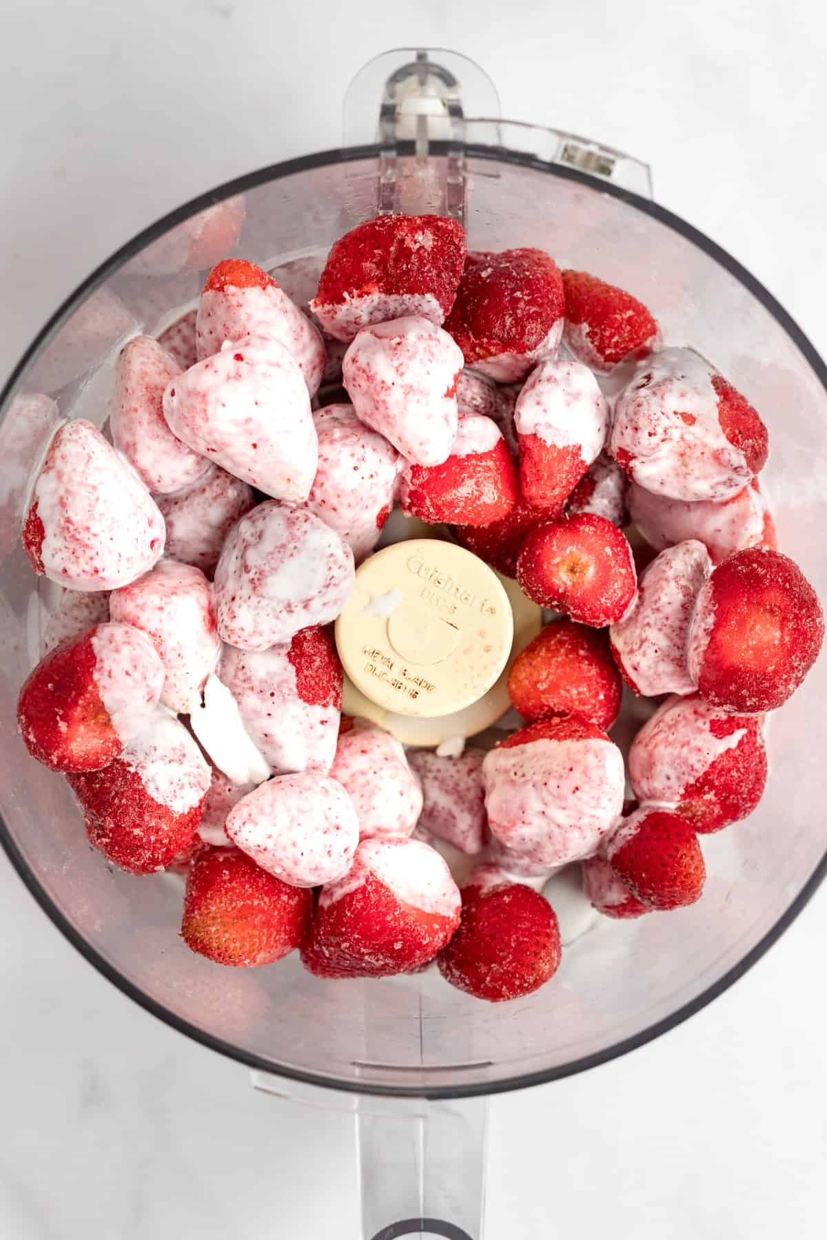 A food processor bowl filled with whole strawberries and scoops of vanilla ice cream, viewed from above.