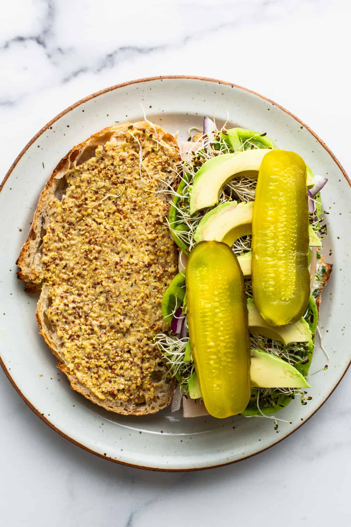 A plate of open-faced sandwich with mustard-covered bread, sprouts, sliced avocado, and two whole pickles on top.