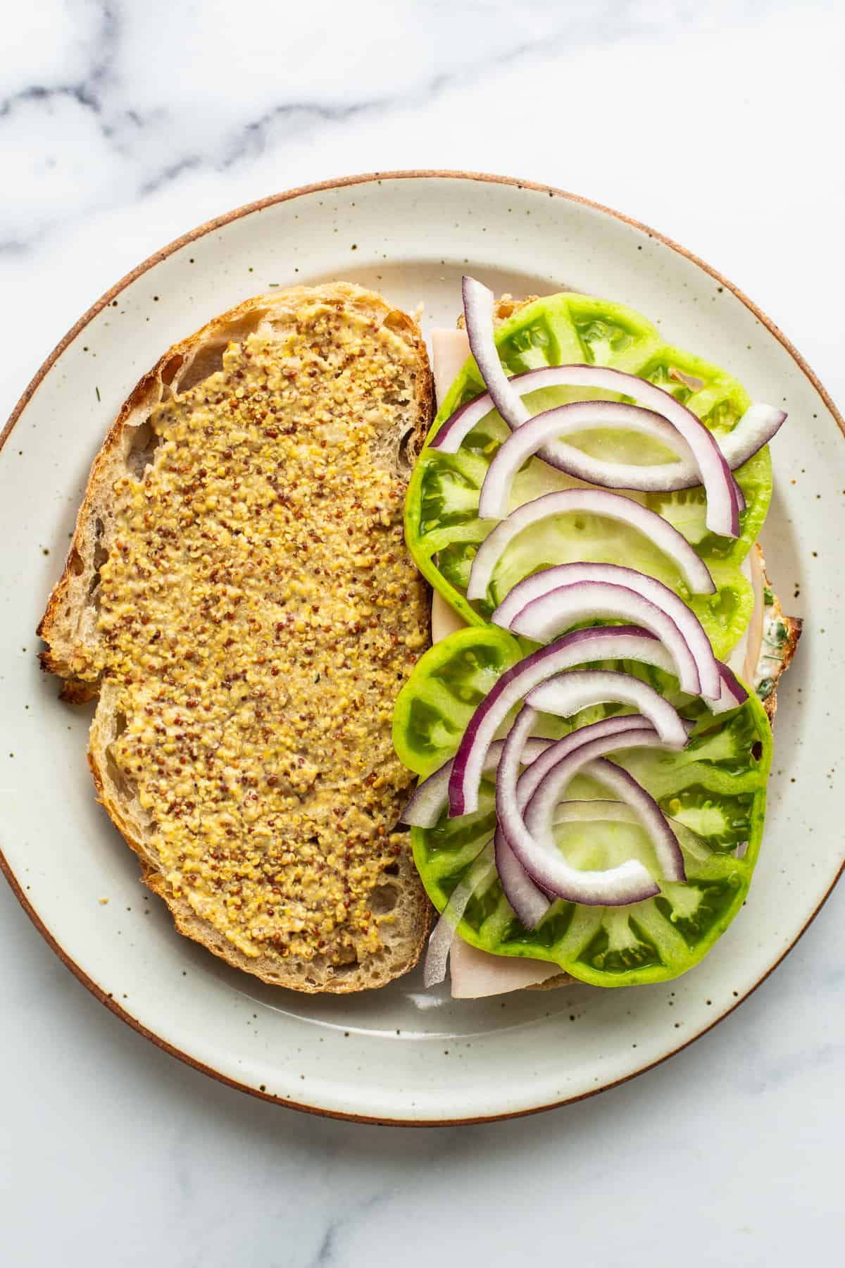 Open-faced sandwich with mustard on whole grain bread, topped with lettuce, cucumber slices, and red onion rings on a ceramic plate.