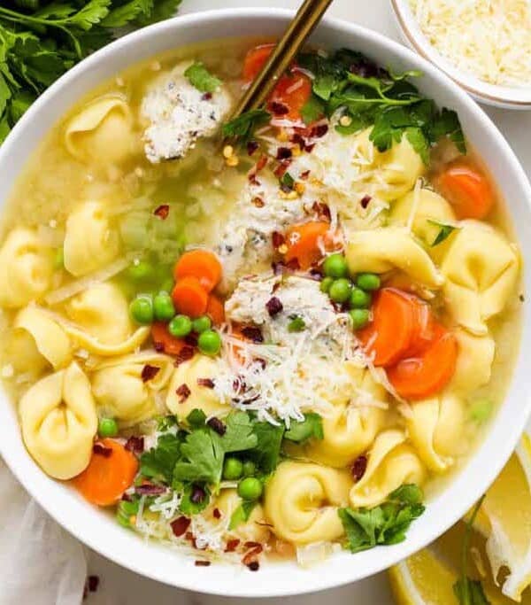 A bowl of tortellini soup with carrots, peas, and garnished with parsley and grated cheese.