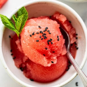 A bowl of watermelon sorbet garnished with mint and black sesame seeds, served with a spoon.