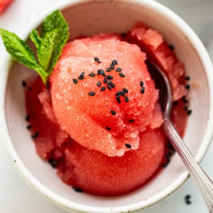 A bowl of watermelon sorbet garnished with mint leaves and black sesame seeds, accompanied by a spoon, on a light surface.