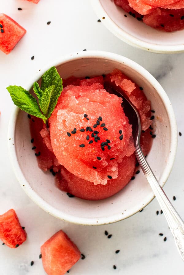 A bowl of watermelon sorbet garnished with mint and black sesame seeds, with a spoon, on a white surface scattered with diced watermelon pieces.