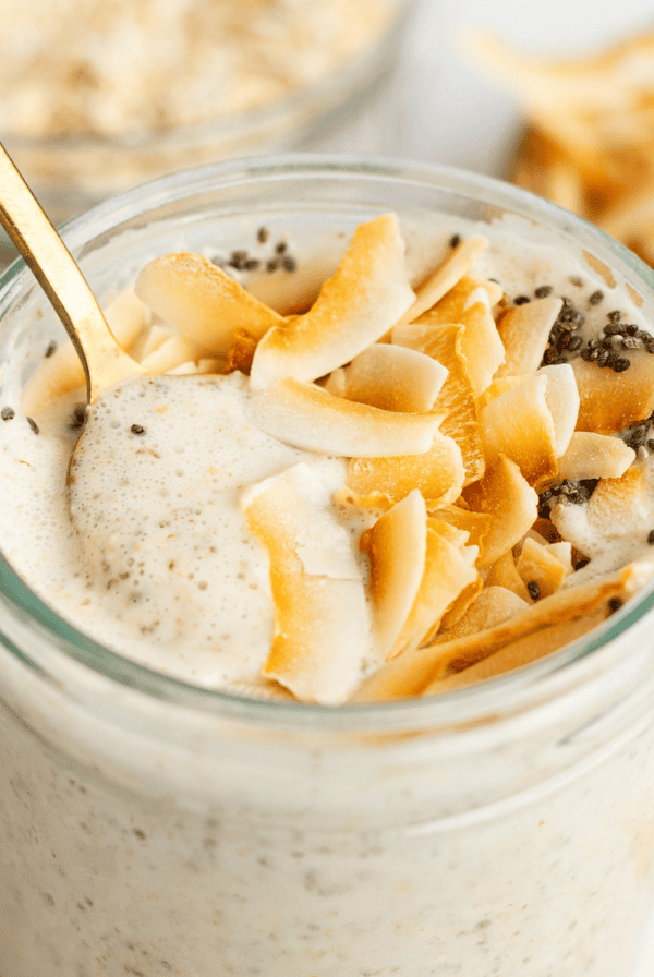 A glass jar filled with creamy coconut latte overnight oats, topped with toasted coconut flakes and chia seeds, with a gold spoon inserted.