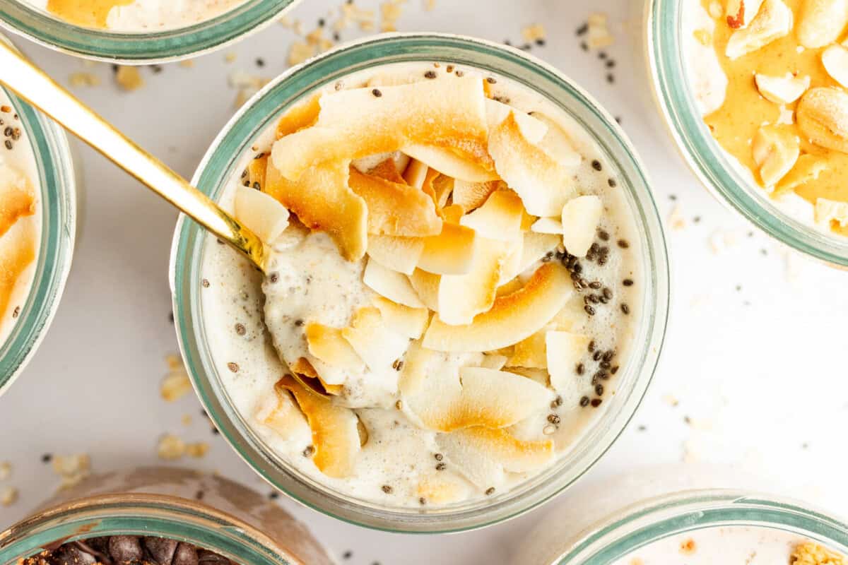 A glass bowl filled with a creamy mixture topped with toasted coconut flakes and chia seeds, with a gold spoon inserted into it. Other similar bowls are partially visible around it.