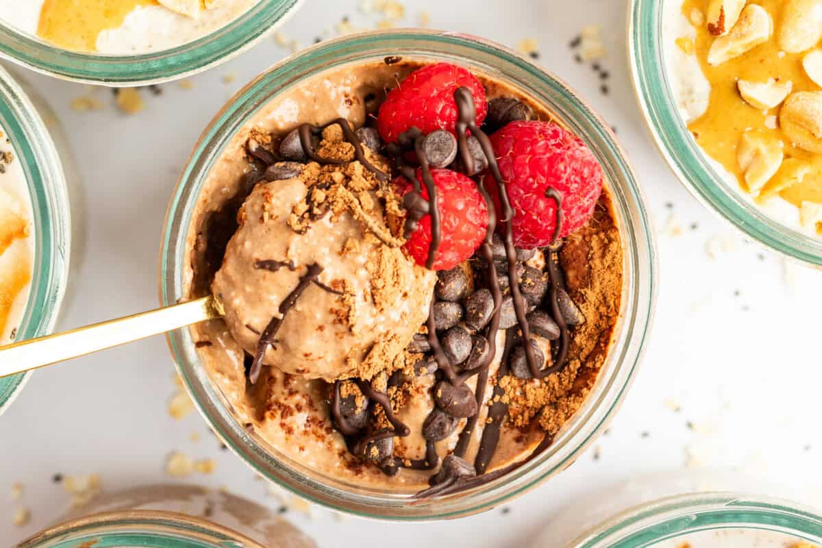 A glass bowl of chocolate oatmeal topped with raspberries, chocolate chips, and a dusting of cocoa powder. A spoon rests inside the bowl. Other oatmeal bowls are partially visible around it.